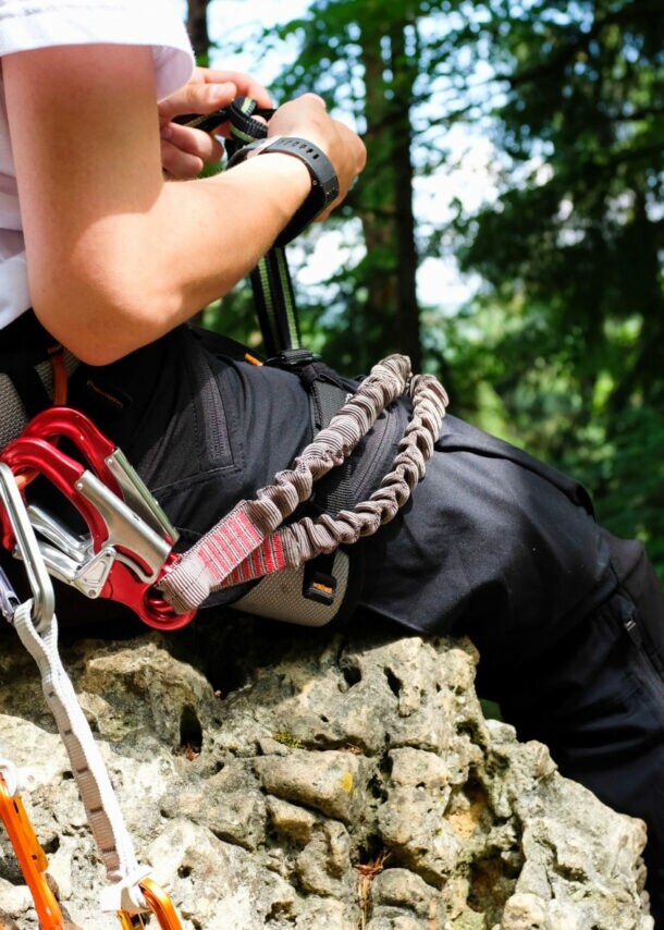 Eine unkenntliche Person sitzt auf einem Felsen im Wald und trägt einen Gürtel um die Hüfte, an dem mehrere Karabiner hängen.