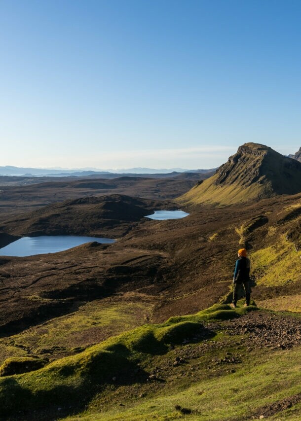 Eine Person steht in einer Landschaft aus Wiesen, Hügeln und Seen in Schottland. 