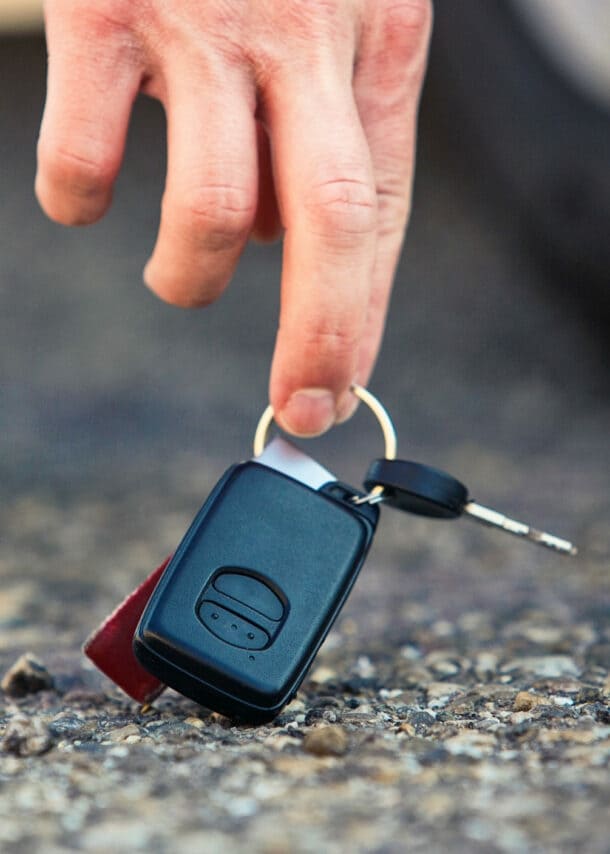 Nahaufnahme einer Hand, die nach einem Autoschlüssel am Asphaltboden greift, dahinter ein Autoreifen und Sneaker in Unschärfe.