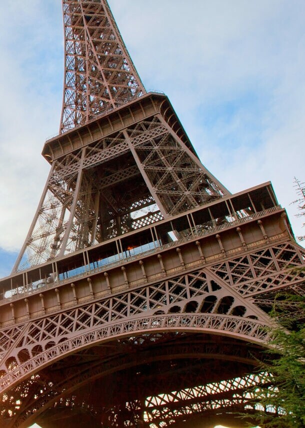 Mittelteil des Eiffelturmes mit Personen auf einer Aussichtsplattform, im Vordergrund grüne Bäume.
