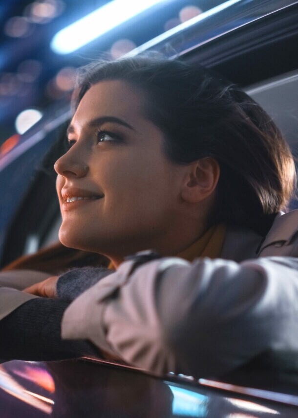 Nahaufnahme einer lächelnden Frau, die von der Rückbank eines fahrenden Autos aus dem geöffneten Fenster blickt, im Hintergrund Leuchtreklame in urbaner Umgebung bei Nacht.