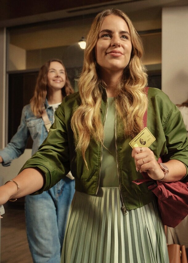Drei junge Frauen gehen fröhlich mit Trolleys durch eine Hotellobby, die Frau vorne hält eine goldene Kreditkarte von American Express in ihrer Hand.