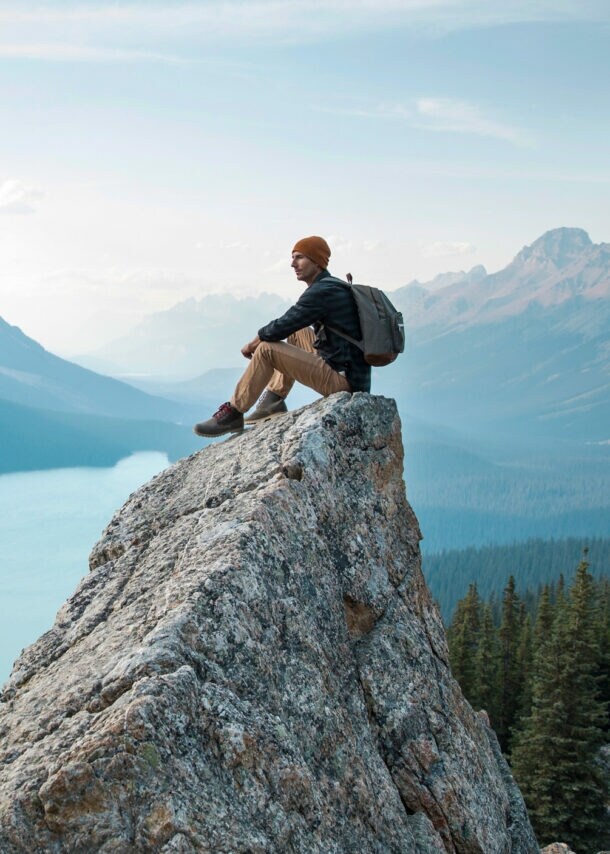 Ein Mann mit Rucksack sitzt auf einem spitzen Felsen in einer Fjordlandschaft mit Nadelwald.