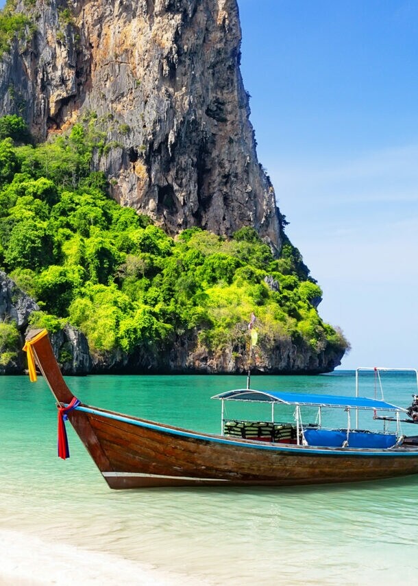 Ein Longtail-Boot im smaragdgrünen Wasser an einem weißen Sandstrand vor einem begrünten Felsen.