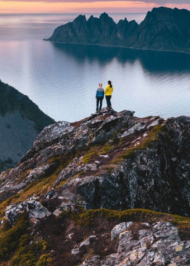 Zwei Personen stehen auf einem Felsen in einer kargen Berglandschaft am Wasser bei Sonnenuntergang.