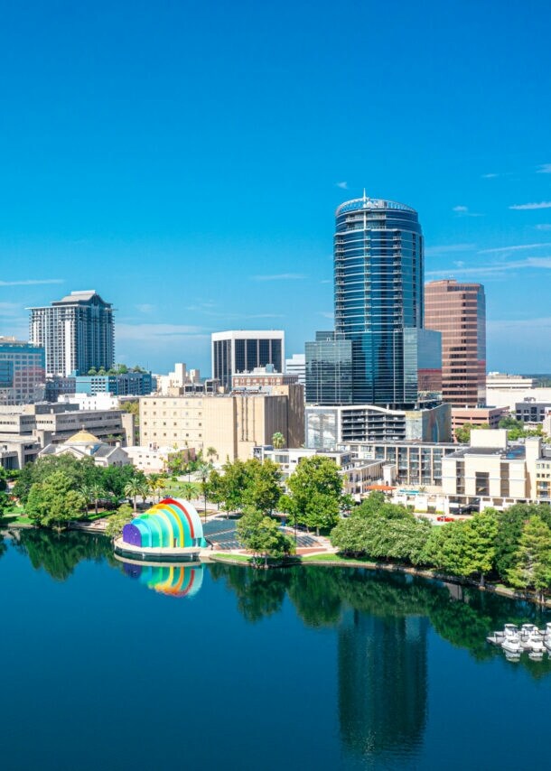 Skyline von Orlando mit Hochhäusern hinter einem See mit Parkanlage unter blauem Himmel.