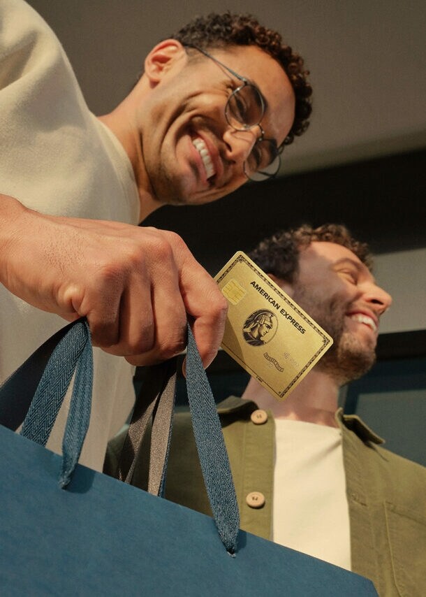 Ein gut gelaunter Mann hält eine goldene Kreditkarte von American Express zusammen mit einer Papiertragetasche in der Hand, daneben ein weiterer Mann.