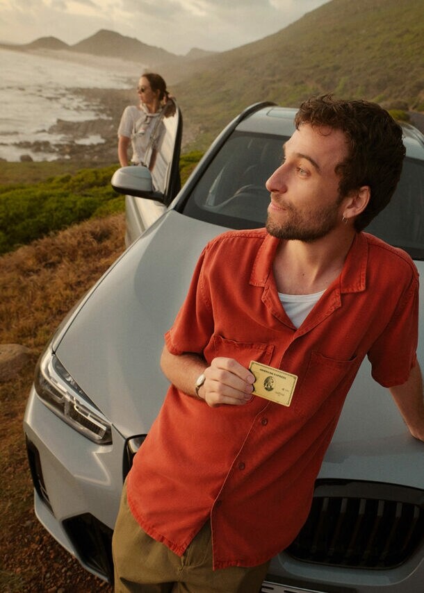 Ein junger Mann lehnt an der Kühlerhaube eines Autos in einer Küstenlandschaft und blickt aufs Meer, in einer Hand hält er eine goldene Kreditkarte von American Express, dahinter zwei weitere Personen am Auto.