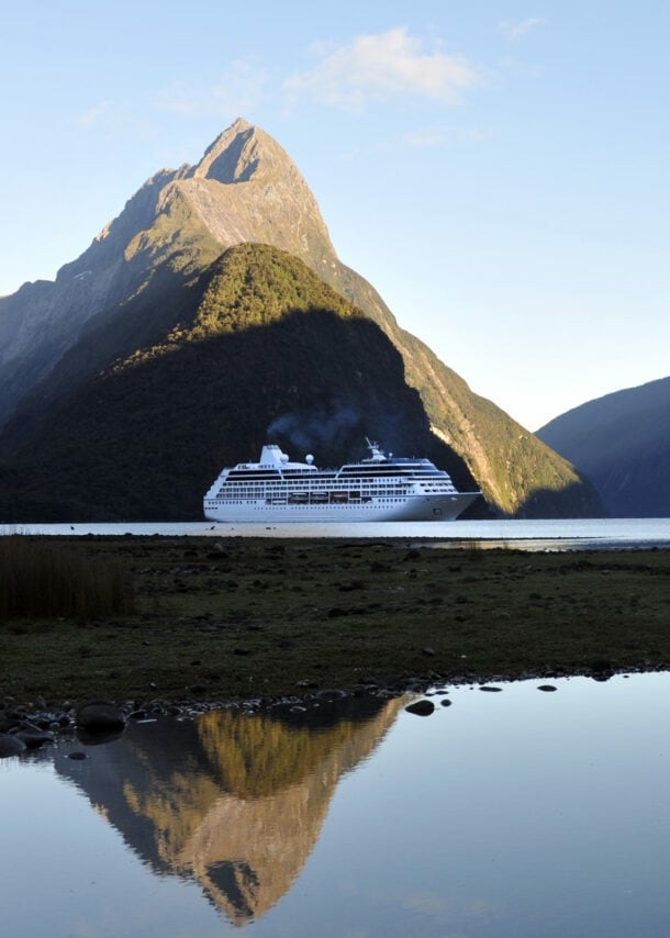 Ein Kreuzfahrtschiff durchquert den Milford Sound in Neuseeland, während sich die hügelige Landschaft des Hintergrunds im ruhigen Wasser spiegelt.