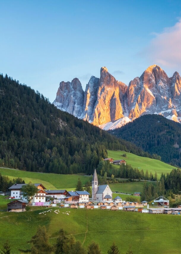 Blick auf das idyllische Dorf Sankt Maddalena, eingebettet zwischen grünen Hügeln, Wäldern und den markanten Geislerspitzen im Hintergrund.