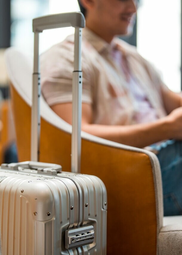 Ein Mann sitzt im Wartebereich eines Flughafens. Im Vordergrund steht sein Rollkoffer.