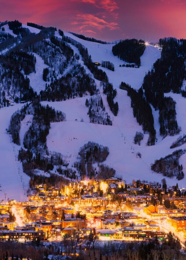 Bergdorf mit beleuchteten Gebäuden und schneebedeckten Skipisten bei Sonnenuntergang.