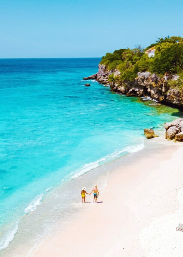Zwei Personen auf einem weißen Sandstrand an türkisblauem Meer in einer Felsbucht mit grüner Vegetation.