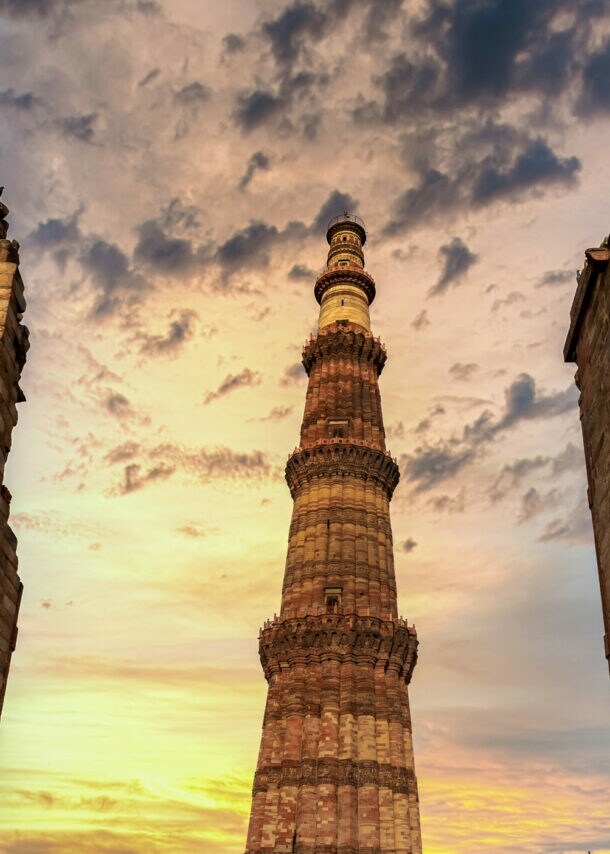 Das Qutub Minar, ein hoher roter Sandsteinturm, umgeben von Ruinenmauern bei Sonnenuntergang mit bewölktem Himmel