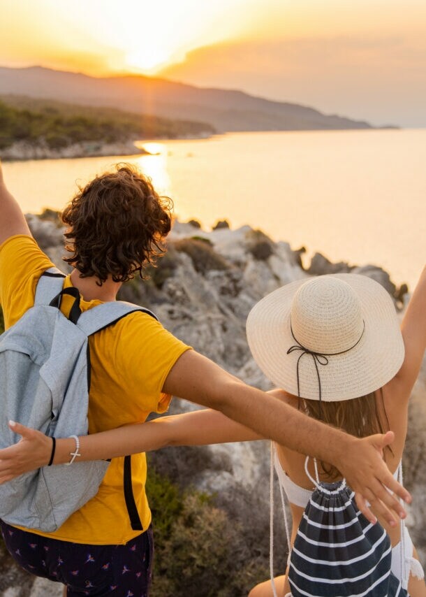 Ein Paar steht mit offenen Armen auf einem Felsen und schaut auf das Meer und den Sonnenuntergang.