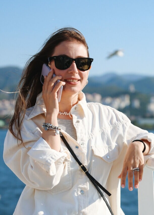 Eine Frau mit Sonnenbrille und wehenden Haaren telefoniert mit einem Smartphone an der Reling eines Schiffes auf dem Meer, im Hintergrund fliegen Möwen vor Stadtpanorama an einer Küste.