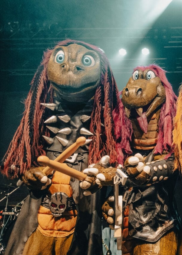 Vier Personen in Dinosaurierkostümen, die in die Kamera gucken und teilweise Instrumente und ein Mikrofon halten