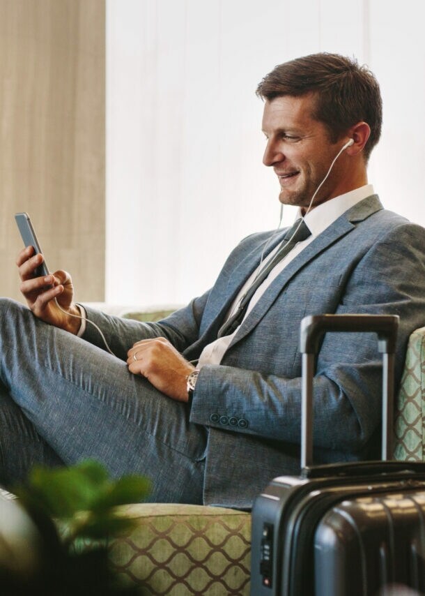 Ein Geschäftsmann sitzt in einem Sessel in einer Flughafen-Lounge und schaut lächelnd auf sein Smartphone.