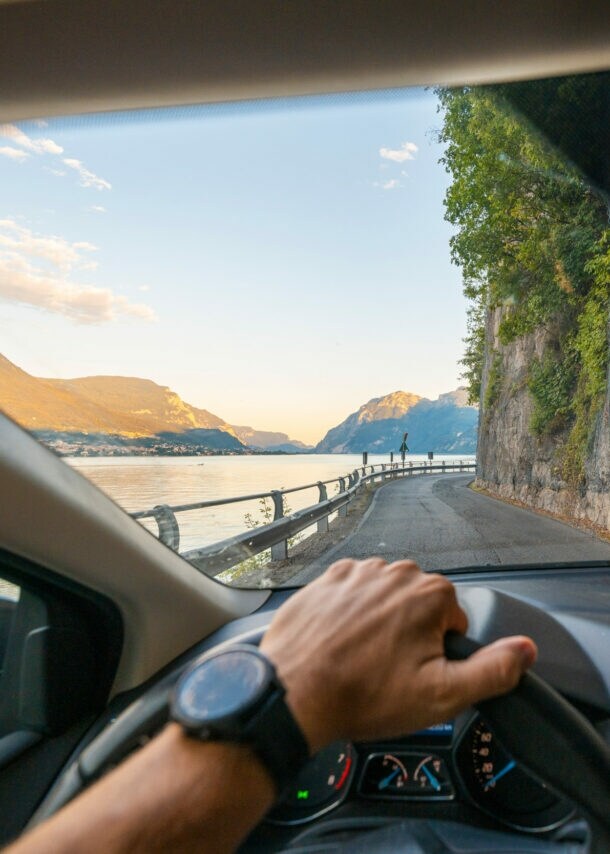 Blick auf eine Küstenstraße entlang einer Felswand durch die Windschutzscheibe eines Autos, im Vordergrund eine Hand am Lenkrad.