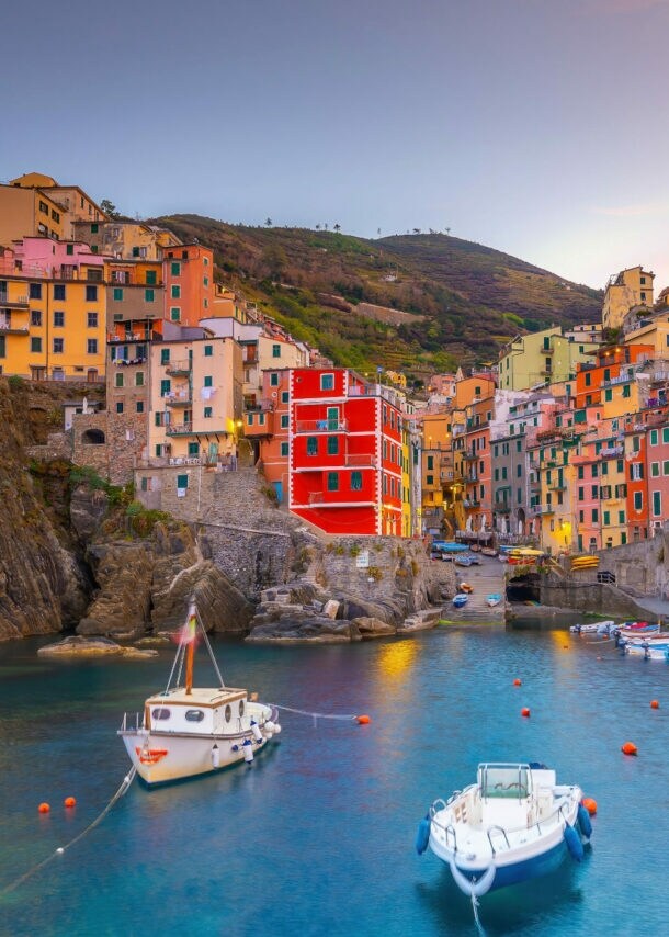 Ortschaft mit bunten Häusern im mediterranen Stil an einer Felsküste mit Meeresbucht mit Fischerbooten im Abendlicht.