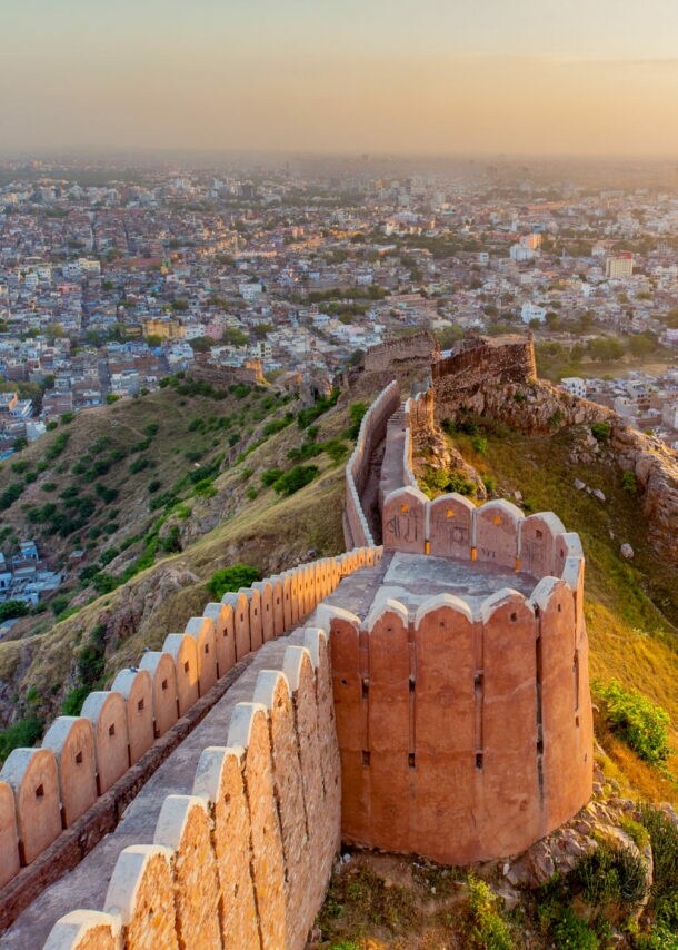 Luftaufnahme der historischen Stadtmauer von Jaipur, die sich über grüne Hügel erstreckt, mit der Stadt im Hintergrund bei Sonnenuntergang