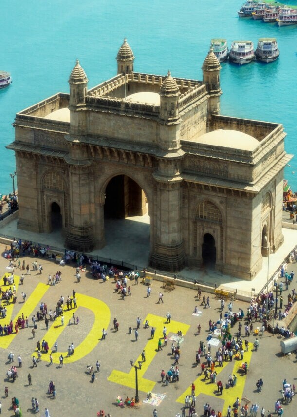 Das Gateway of India, ein großes Tor mit Bögen und Türmen, umgeben von Menschen und Booten im Wasser in Mumbai.