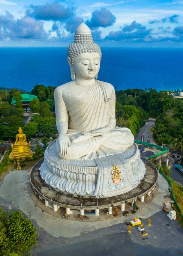Eine große weiße Buddha-Statue auf einem bewaldeten Hügel an einer Meeresküste.