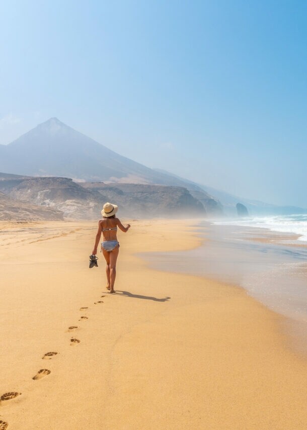 Rückansicht einer Frau im Bikini, die einen breiten Sandstrand entlangläuft, im Hintergrund spitze Felsformationen.