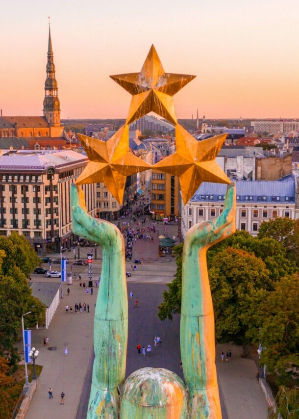 Stadtpanorama von Riga mit Altstadt aus der Luft, im Vordergrund die Arme einer Freiheitsstatue mit drei Sternen.