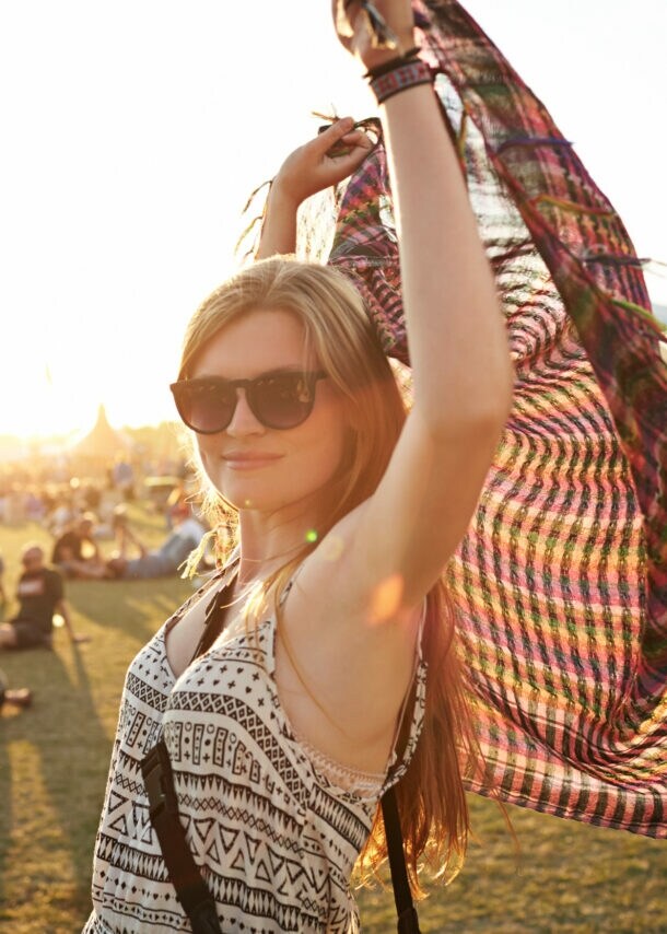 Eine Frau mit Sonnenbrille steht auf einer Wiese, auf der Leute sitzen, und hält ein Tuch über ihren Kopf.