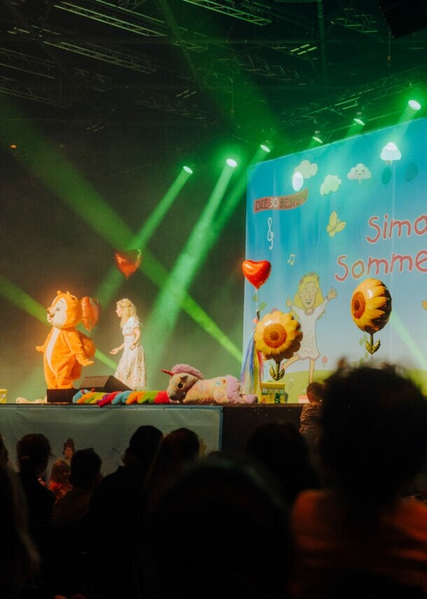 Simone Sommerland steht auf einer Bühne voller Luftballons, neben ihr eine Person im Eichhörnchen-Kostüm, vor ihr Publikum.