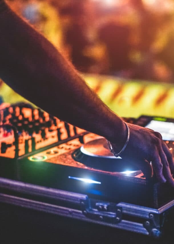 Ein DJ steht vor seinem Mischpult, im Hintergrund ist verschwommen sein Publikum zu sehen.