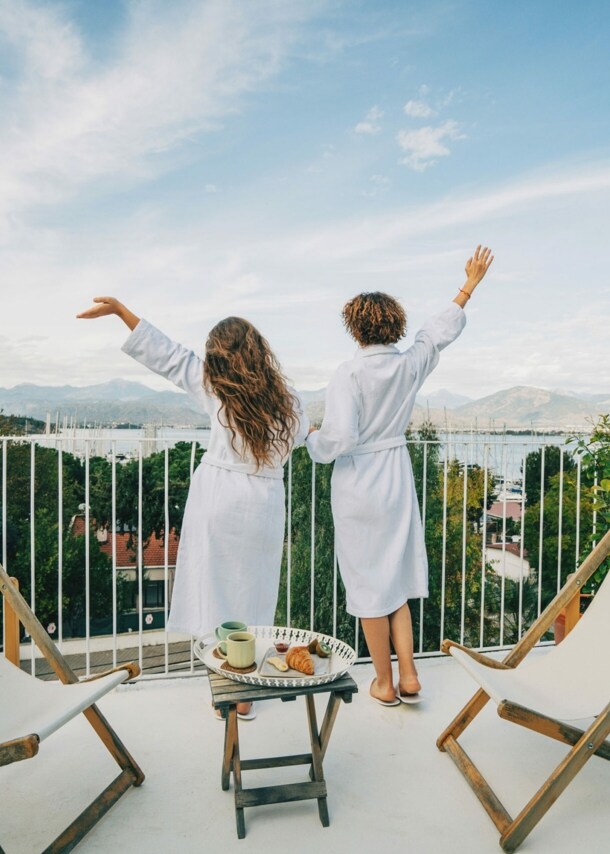 Zwei Menschen in weißen Bademänteln von hinten stehen auf einem Balkon, blicken ins Grüne und strecken freudevoll die Arme in die Luft.