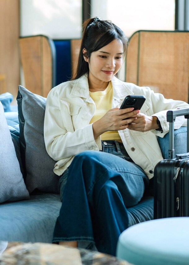 Eine junge asiatische Frau sitzt iauf einem Sessel in einer Lounge und schaut auf ihr Smartphone, neben ihr steht ein Hartschalenkoffer.