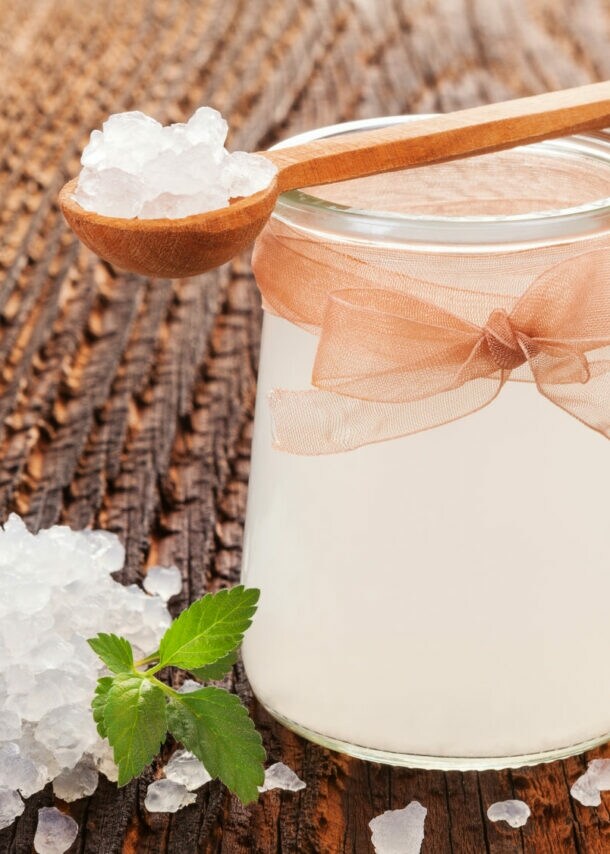 Glas mit milchiger Flüssigkeit und Schleife, umgeben von kristallartigen weißen Körnern auf Holzoberfläche