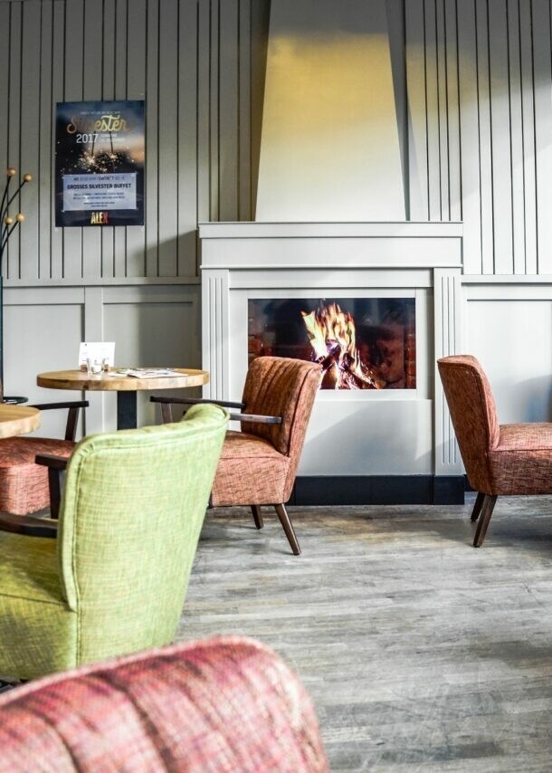 Gemütlicher Gastraum des ALEX in Wiesbaden mit loderndem Gaskamin, runden kleinen Tischen und bequemen Sesseln in Grün und Rot.