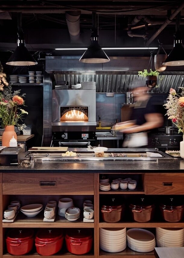 Blick in die Küche, wo im Ofen ein Feuer brennt und neben der unverputzten Backsteinwand eine Anrichte mit Blumensträußen und frischen Kräutern steht.