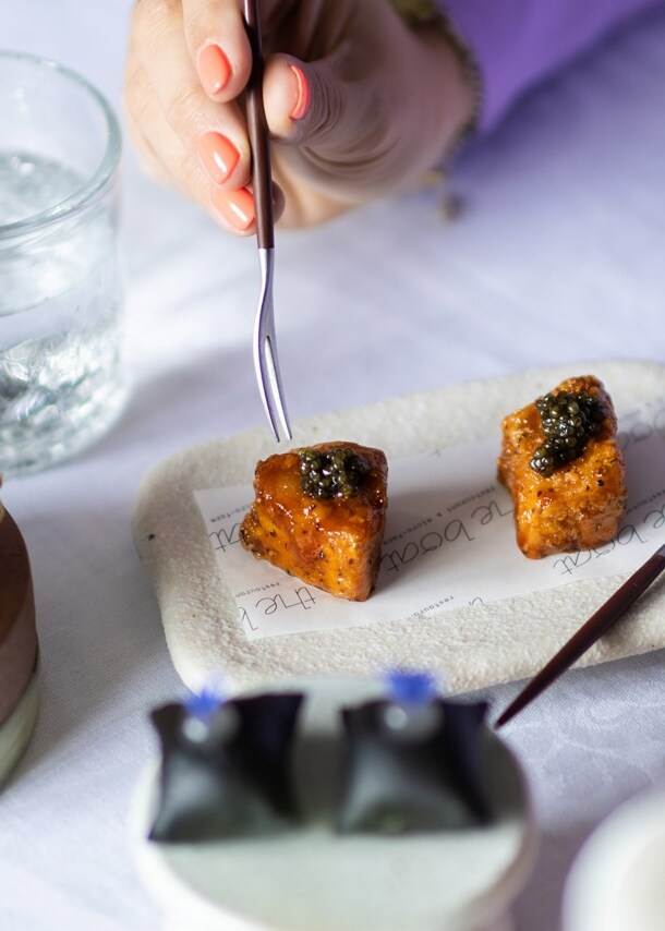 Eine Hand führt eine Gabel zu einem kunstvoll angerichteten Dessert auf einem weiß eingedeckten Restauranttisch.