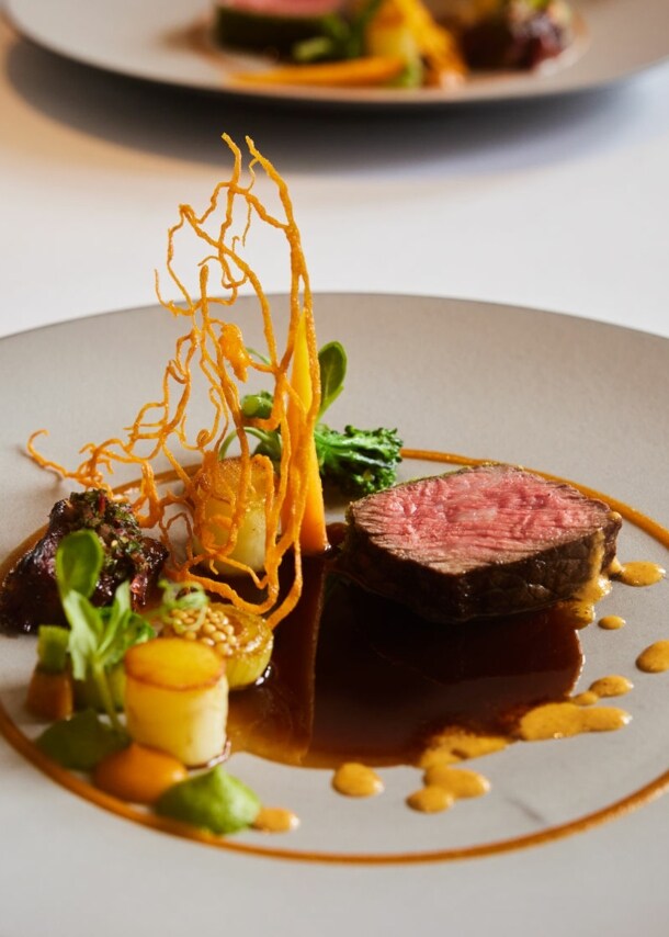 Kunstvoll arrangiertes Gericht mit einem Stück Fleisch auf einem großen weißen Teller auf einem weißen Tisch.