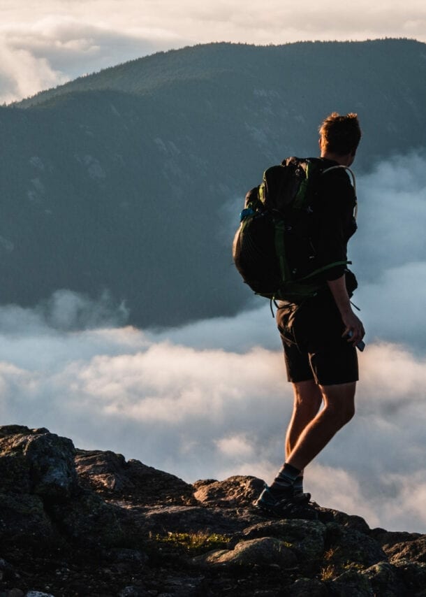 Wanderer mit Rucksack steht auf felsigem Gipfel und blickt auf nebelverhangene Berge bei Sonnenuntergang