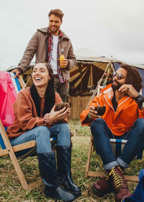 Drei Personen sitzen und stehen entspannt auf einem Festivalgelände mit Zelten im Hintergrund, lachen und trinken Getränke.