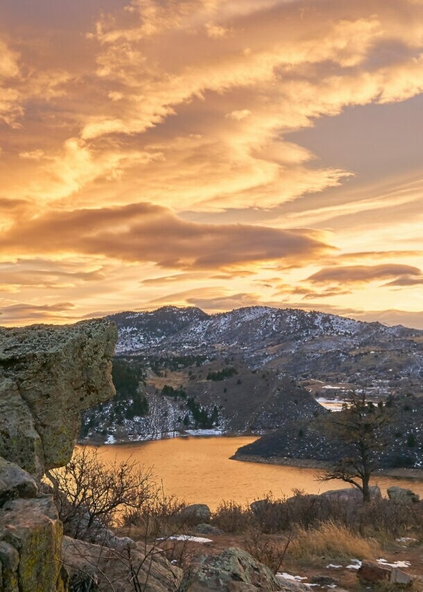 Felsige Landschaft mit Nadelbäumen und See bei Sonnenuntergang unter bewölktem Himmel