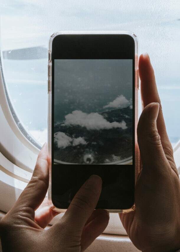 Hände halten ein Smartphone vor einem Flugzeugfenster, das den Himmel mit Wolken zeigt.