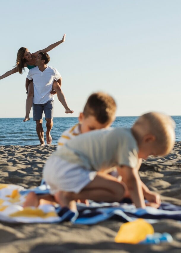 Zwei spielende Kinder auf einem Strandtuch am Strand, im Hintergrund trägt ein Mann eine Frau auf dem Rücken am Meer