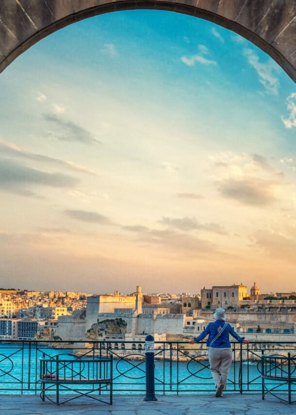 Blick durch einen großen Steinbogen auf eine Stadt am Wasser bei Sonnenuntergang, eine Person lehnt am Geländer