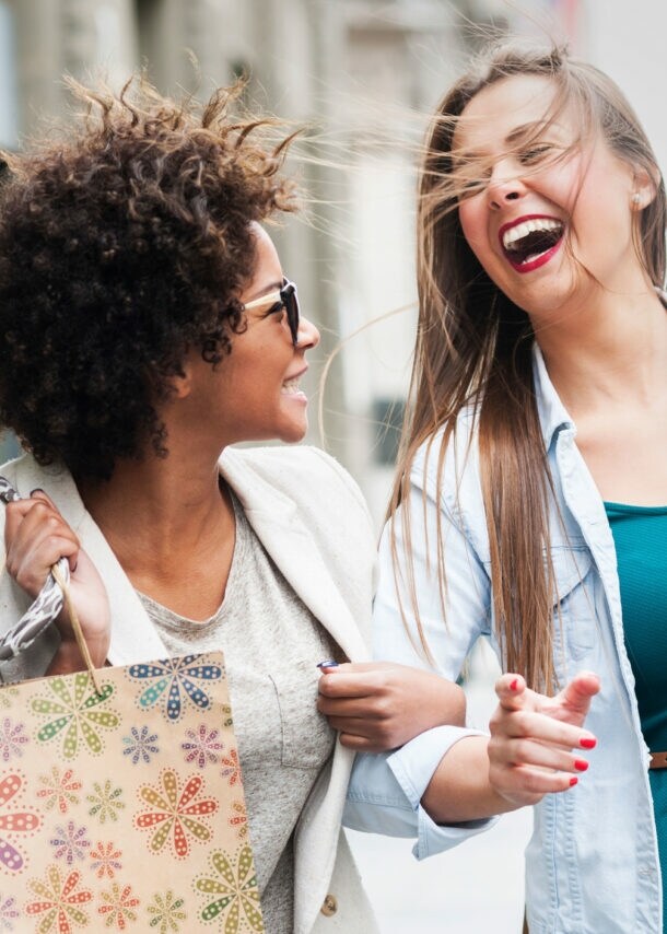 Zwei lachende Frauen mit Einkaufstüten auf dem Gehweg einer Straße.
