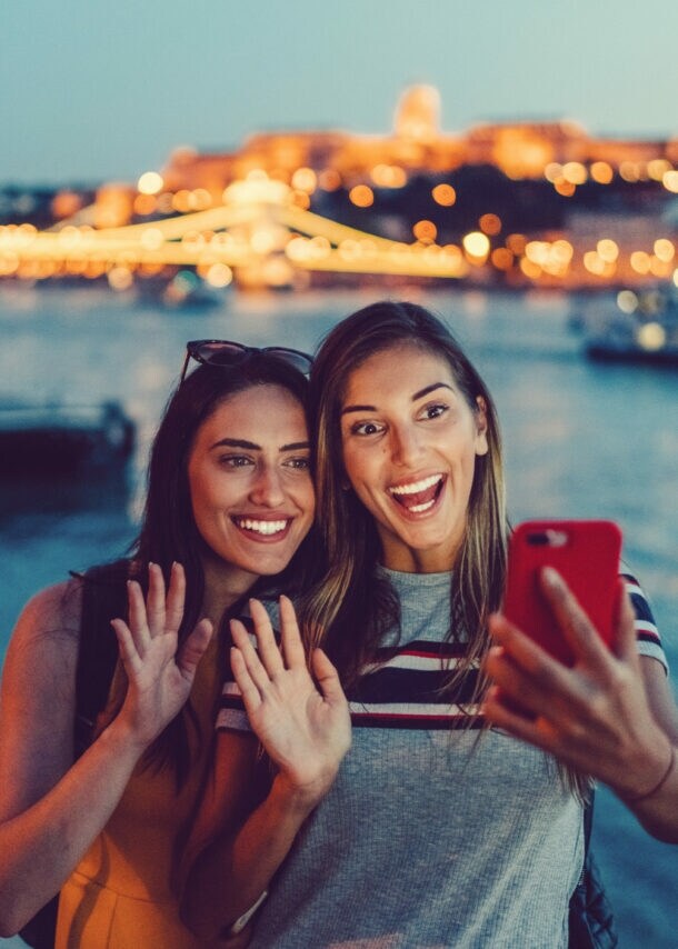 Zwei junge Frauen machen abends am Flussufer ein Selfie, im Hintergrund beleuchtete Brücke und Stadtlichter
