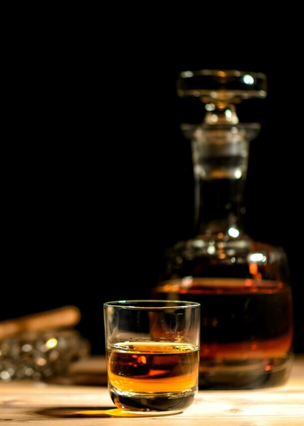 Beiger Strohhut mit schwarzem Band neben einem Glas und einer Karaffe mit braunem Getränk auf einem Holztisch vor schwarzem Hintergrund