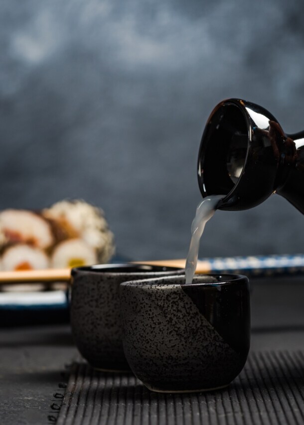 Hand gießt Sake aus einer schwarzen Karaffe in eine kleine schwarze Tasse, im Hintergrund Sushi und Essstäbchen