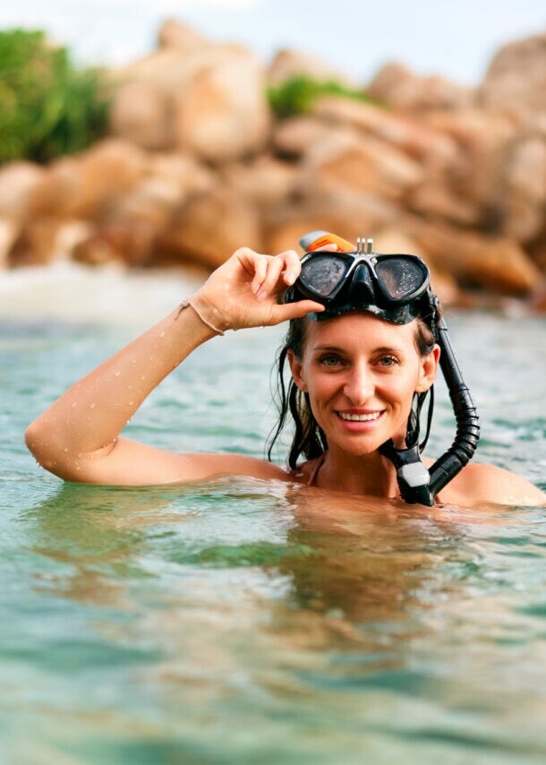 Frau mit Schnorchelmaske im Wasser vor felsiger Küste, hebt die Maske lächelnd an